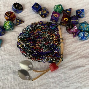 May include: A collection of polyhedral dice in various colors, including blue, green, and purple, scattered around a chainmail dice bag. The bag is made of colorful metal rings and has a leather strap and a red toggle. Two silver scale-shaped charms are also present.