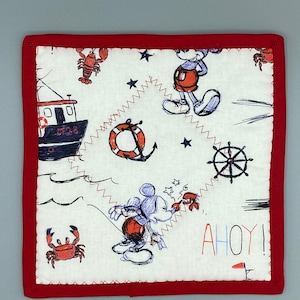 May include: Square, white fabric item with a red border, featuring nautical-themed illustrations. Mickey Mouse is depicted with crabs, a boat, and a ship's wheel. The word "AHOY!" is written in colourful letters. The item is likely a decorative piece or a kitchen item.