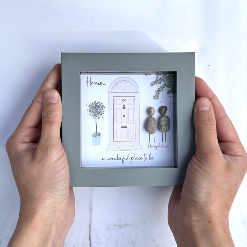 Puede incluir: Una ilustraci&oacute;n enmarcada de una casa con una puerta rosa y un &aacute;rbol en maceta. Dos personas de guijarros se paran frente a la casa con el texto "Harry & Lisa, un lugar maravilloso para estar."