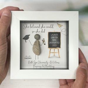 May include: A square white framed wall art piece featuring a pebble art figure. The text reads: She believed she could, so she did. A chalkboard on an easel says: LAUREN CARTER HAS GRADUATED 2023. Below is: Bath Spa University - BA Hons Business & Marketing.