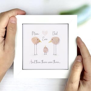 Op de afbeelding: Een ingelijste kunstdruk met een witte rand. Het kunstwerk toont drie vogelachtige figuren gemaakt van textuurmateriaal, met de woorden "Mum", "Dad" en "Evie" erboven. De zin "And then there were three..." staat eronder.