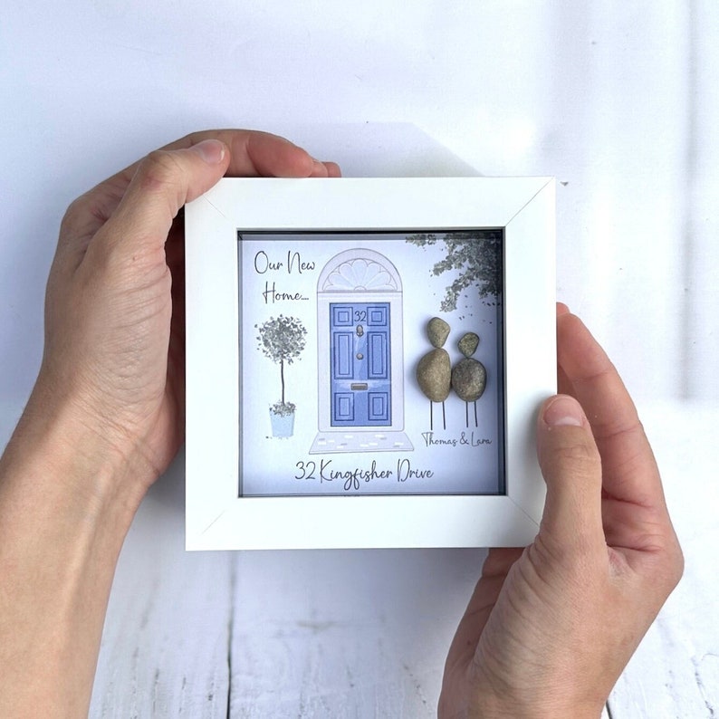 Puede incluir: Una imagen enmarcada en blanco con una puerta azul y dos personas de guijarros de pie frente a ella. El texto dice "Nuestro nuevo hogar... Thomas y Laura 32 Kingfisher Drive."