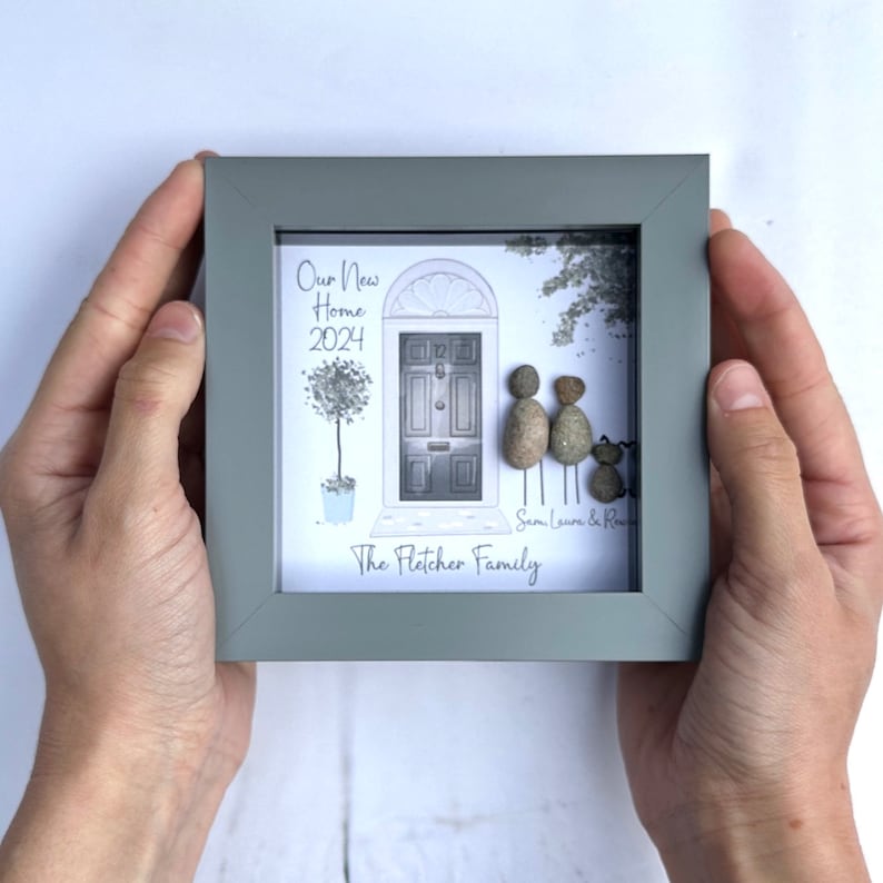 Puede incluir: Una pieza de arte de guijarros enmarcada que representa una casa con una puerta, dos figuras y un perro. El texto dice "Our New Home 2024" y "The Fletcher Family".