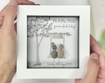 Könnte beinhalten: Ein weiß gerahmtes Bild mit einem Kieselstein-Kunstdesign eines Paares, das auf einer Schaukel unter einem Baum sitzt. Das Schild über der Schaukel lautet "Just Married". Der Text unter dem Paar lautet "Mr & Mrs Fletcher forever starts today! Saturday 26th August 2023."