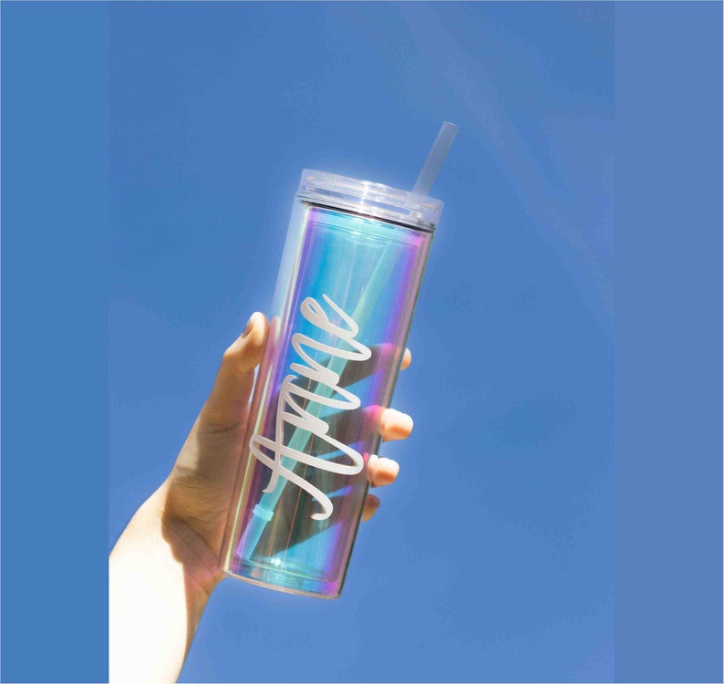 May include: A clear iridescent tumbler with a white straw and the name "Anne" written on the side. The tumbler is being held by a hand against a blue sky background.