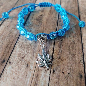 Könnte beinhalten: Ein blaues Kordelarmband mit facettierten, durchscheinenden blauen Perlen und einer zentralen silberfarbenen Metallperle. Ein silberfarbener Metallanhänger in Form eines siebenblättrigen Blattes hängt in der Mitte. Das Armband hat verstellbare Enden.