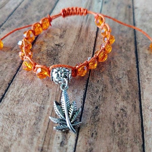 Könnte beinhalten: Ein verstellbares Armband aus orangefarbener Kordel mit facettierten orangefarbenen Kristallperlen und einem silberfarbenen Metallanhänger in Form eines Cannabisblattes. Das Armband hat einen Schiebeknotenverschluss und zwei kleine Perlen.