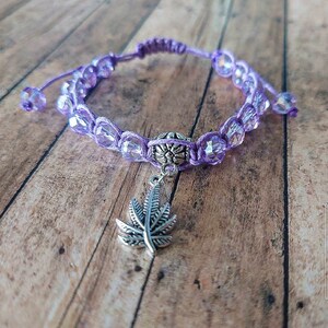 Könnte beinhalten: Ein violettes Kordelarmband mit facettierten, durchscheinenden violetten Perlen und einer zentralen silberfarbenen Metallperle. Ein silberfarbener blattförmiger Anhänger hängt in der Mitte. Das Armband hat einen verstellbaren Verschluss mit zwei kleinen Perlen.