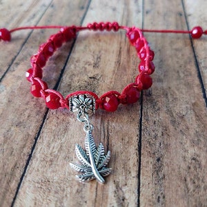 Könnte beinhalten: Ein rotes Kordelarmband mit facettierten roten Perlen und einem silberfarbenen Metallanhänger in Form eines Cannabisblattes. Das Armband verfügt über eine zentrale silberfarbene Zierperle und einen verstellbaren Schiebeknoten mit kleinen roten Perlen.
