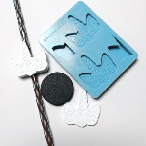May include: A blue silicone mould with two unicorn head designs. The mould is labelled "AMAZING MOLD". There are three white unicorn head charms, one of which is on a brown and white striped straw.