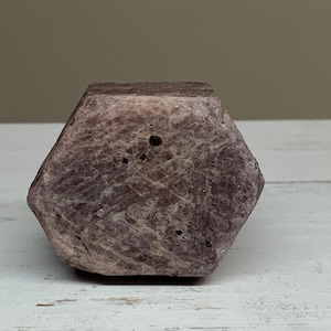 May include: A faceted, natural gemstone with a muted purple hue. The stone has a rough, textured surface and a geometric shape. The gemstone is displayed on a white wooden surface.