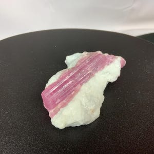 May include: A raw, natural tourmaline gemstone. The stone features a vibrant pink center, encased in a white matrix. The crystal structure is visible, showcasing the mineral's natural form. The gemstone is a unique piece for collectors or jewelry making.