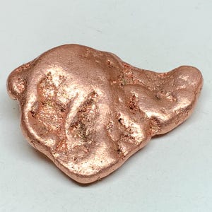 May include: A raw copper nugget with a textured surface and a natural, irregular shape. The nugget has a reddish-brown color and sits on a white surface. The copper appears to have a slightly metallic sheen.