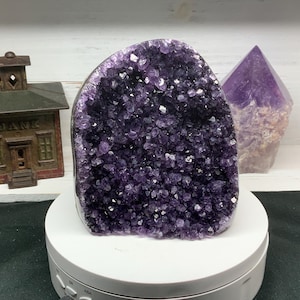 May include: A large, deep purple amethyst geode with sparkling crystal formations. A smaller, polished amethyst point and a vintage metal bank are in the background. The geode sits on a white rotating display stand.