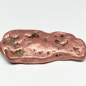 May include: A raw, metallic copper nugget with an irregular, elongated shape. The surface features a textured, reddish-brown appearance with small, deep crevices and dark, oxidized mineral inclusions scattered across its uneven top.