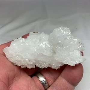 May include: A cluster of white quartz crystals with a sparkling, crystalline structure. The quartz formation has a rough, uneven texture and is held in a hand. The crystals are translucent and reflect light, creating a shimmering effect. The overall shape is irregular and natural.