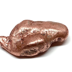 May include: A raw copper nugget with a metallic, reddish-brown luster. The surface of the mineral specimen is irregular and textured, featuring natural indentations and smooth, rounded edges across its elongated, solid form.