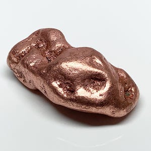 May include: A raw, metallic copper nugget with an irregular, elongated shape. The surface features a textured, pitted appearance with a warm, reddish-brown metallic luster and reflective highlights across its uneven, bumpy contours.