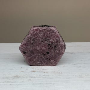 May include: A faceted, hexagonal ruby gemstone. The gem is a deep pinkish-red color with a textured surface and dark inclusions. It sits on a white wooden surface against a neutral background.