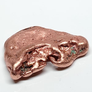 May include: A raw copper nugget with a metallic, reddish-brown surface. The object features an irregular, organic shape with a textured, pitted exterior and small patches of green oxidation visible within the crevices of the metal.