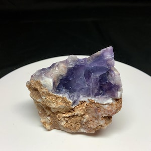 May include: A raw, natural fluorite specimen with a striking combination of colors. The top portion displays a vibrant, translucent purple hue, while the base features a rough, earthy brown and beige matrix. This unique mineral piece is ideal for collectors.