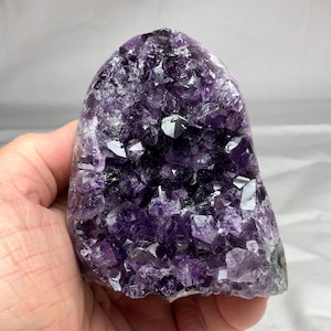 May include: A raw amethyst geode cluster featuring deep purple, multifaceted crystal points. The stone has a rounded, arch-like shape with a hollow center filled with jagged, translucent violet crystals that catch the light from various angles.