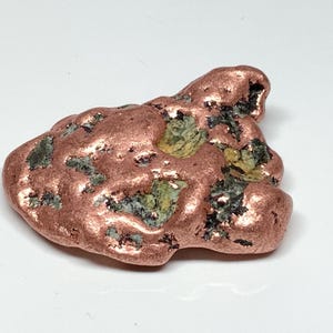 May include: A raw copper nugget featuring a metallic, reddish-brown surface with irregular, smooth, and rounded protrusions. The specimen displays embedded patches of dark grey and pale yellow mineral inclusions within its textured form.