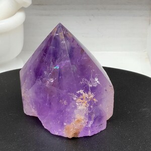 May include: A large, faceted amethyst crystal point. The crystal is a deep purple color with some areas of lighter purple and brown. It sits on a black surface, with a white mortar and pestle in the background.