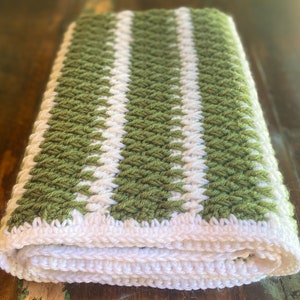 May include: A green and white striped crocheted blanket folded on a wooden surface. The blanket is made with a textured stitch and has a white border.