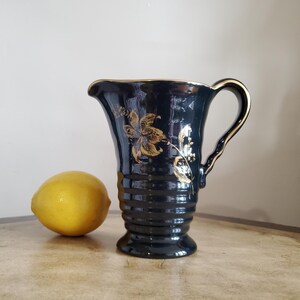 May include: A black ceramic pitcher with a gold floral design and a ribbed body. The pitcher has a handle and a wide mouth.