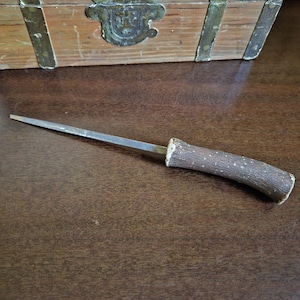 May include: A handcrafted knife with a wooden handle and a silver blade. The handle is made from a piece of wood with a natural, textured appearance. The blade is long and slender, with a sharp edge.