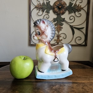 May include: A vintage ceramic rocking horse planter in white, brown, and gray, with a yellow saddle and blue base. An apple is next to the planter. The planter has a detailed design and is likely a decorative item.
