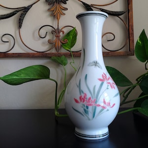 May include: White ceramic vase with a flared top and a round base. The vase is decorated with pink flowers, green stems, and a butterfly. Black bands encircle the top and base. The vase is approximately 8 inches tall.