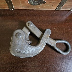 May include: A weathered, silver-colored cable grip tool. The tool has a curved hook shape with a hinged lever and a loop handle. The text "1604-201" is visible on the tool's surface. The tool is designed for gripping and securing cables.
