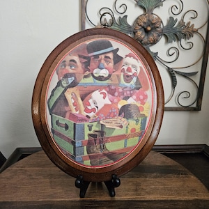 May include: An oval wooden wall plaque featuring a printed illustration of several clowns emerging from a green trunk. The scene includes a makeup box, a pair of boots, and various clown faces with colorful makeup, all framed by a dark wood border.