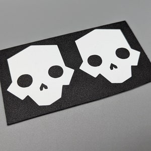 May include: Two white geometric skull stickers on a black rectangular background. Each skull has large circular eye sockets and a small nose cavity. The stickers are likely for decoration or customization.