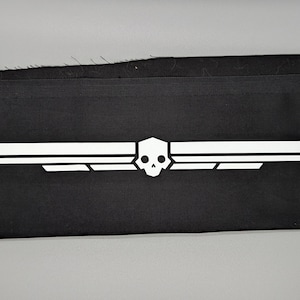 Pode incluir: Um patch de tecido preto com um emblema branco. O emblema é um design simétrico com uma caveira no centro, ladeada por formas de asa. O patch é retangular e parece ser feito de um material durável.