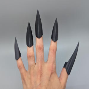 May include: A hand with five black, pointed claw-like finger covers. The claw covers are made of a matte black material and are attached to the fingers. The background is a solid, light grey colour. The claws are a unique accessory.