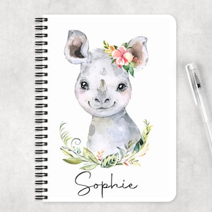 May include: A white spiral bound notebook featuring a watercolour illustration of a grey baby rhinoceros wearing a floral crown. The name "Sophie" is written in black cursive at the bottom of the notebook.