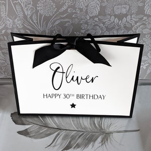 May include: A white gift bag with black trim and a black bow. The bag has the name "Oliver" and the text "HAPPY 30TH BIRTHDAY" printed on it, along with a small black star. The background is a gray floral wallpaper.