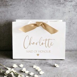 K&ouml;nnte beinhalten: Eine wei&szlig;e Geschenkt&uuml;te mit einer goldenen Satinschleife an der Oberseite. Auf der Vorderseite ist der Name Charlotte in goldener Schreibschrift aufgedruckt, gefolgt von dem Text MAID OF HONOUR in goldenen Gro&szlig;buchstaben und einem kleinen Herz.