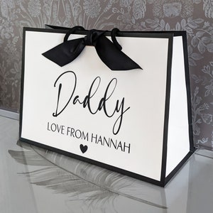 May include: A white gift bag with a black border and a black satin ribbon. The bag has the text "Daddy Love From Hannah" printed on the front.