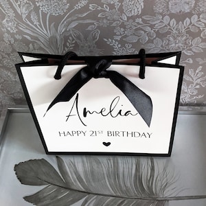 May include: White gift bag with black trim and handles, tied with a black ribbon. The bag features the name "Amelia" and the text "HAPPY 21ST BIRTHDAY" in black script. A small black heart is below the text.