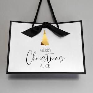May include: White gift bag with a black border and handles, featuring a gold Christmas tree design and the text "MERRY Christmas ALICE" in black script. A black bow is tied at the top.