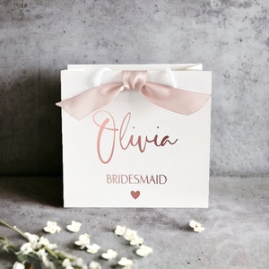 K&ouml;nnte beinhalten: Eine quadratische wei&szlig;e Geschenkt&uuml;te mit einem Griff aus altrosa Satinband. Auf der Vorderseite ist der Name Olivia in einer metallisch ros&eacute;goldenen Schreibschrift gedruckt, gefolgt von dem Wort BRIDESMAID in Gro&szlig;buchstaben und einem kleinen Herz.