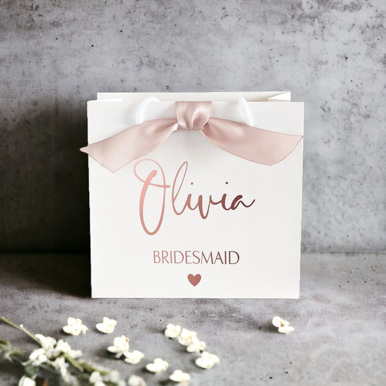 K&ouml;nnte beinhalten: Eine quadratische wei&szlig;e Geschenkt&uuml;te mit einem Griff aus altrosa Satinband. Auf der Vorderseite ist der Name Olivia in einer metallisch ros&eacute;goldenen Schreibschrift gedruckt, gefolgt von dem Wort BRIDESMAID in Gro&szlig;buchstaben und einem kleinen Herz.