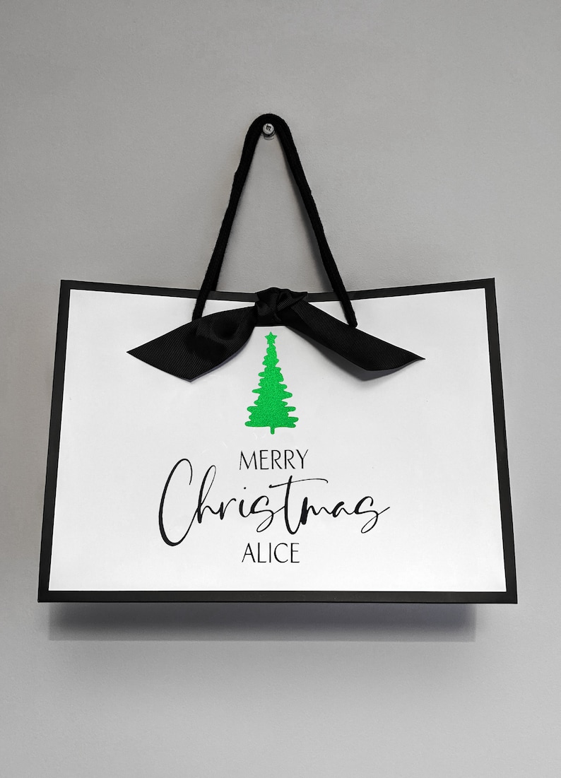 May include: White gift bag with a black border, black ribbon handles, and a black bow. The bag features a green Christmas tree graphic and the text "MERRY Christmas ALICE" in black script.