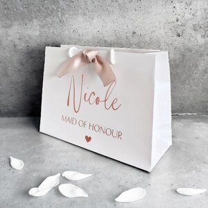 K&ouml;nnte beinhalten: Eine wei&szlig;e Geschenkt&uuml;te mit einem altrosa Schleifenband und dem Namen Nicole in Schreibschrift &uuml;ber dem Text MAID OF HONOUR und einem kleinen Herzsymbol. Die T&uuml;te hat wei&szlig;e Kunststoffkordelgriffe und eine klare, rechteckige Struktur.