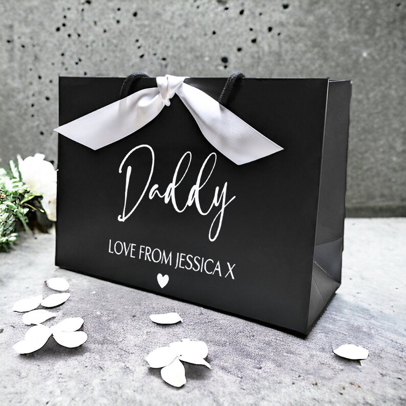 May include: Black gift bag with a white satin ribbon and the text "Daddy Love from Jessica X" in white lettering.