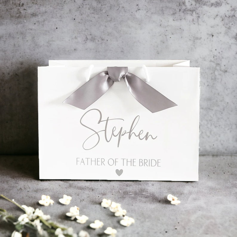 K&ouml;nnte beinhalten: Eine wei&szlig;e Geschenkt&uuml;te mit einer grauen Satinschleife an der Oberseite. Auf der Vorderseite ist der Name Stephen in einer Schreibschrift aufgedruckt, gefolgt von dem Text FATHER OF THE BRIDE und einem kleinen grauen Herzsymbol darunter.